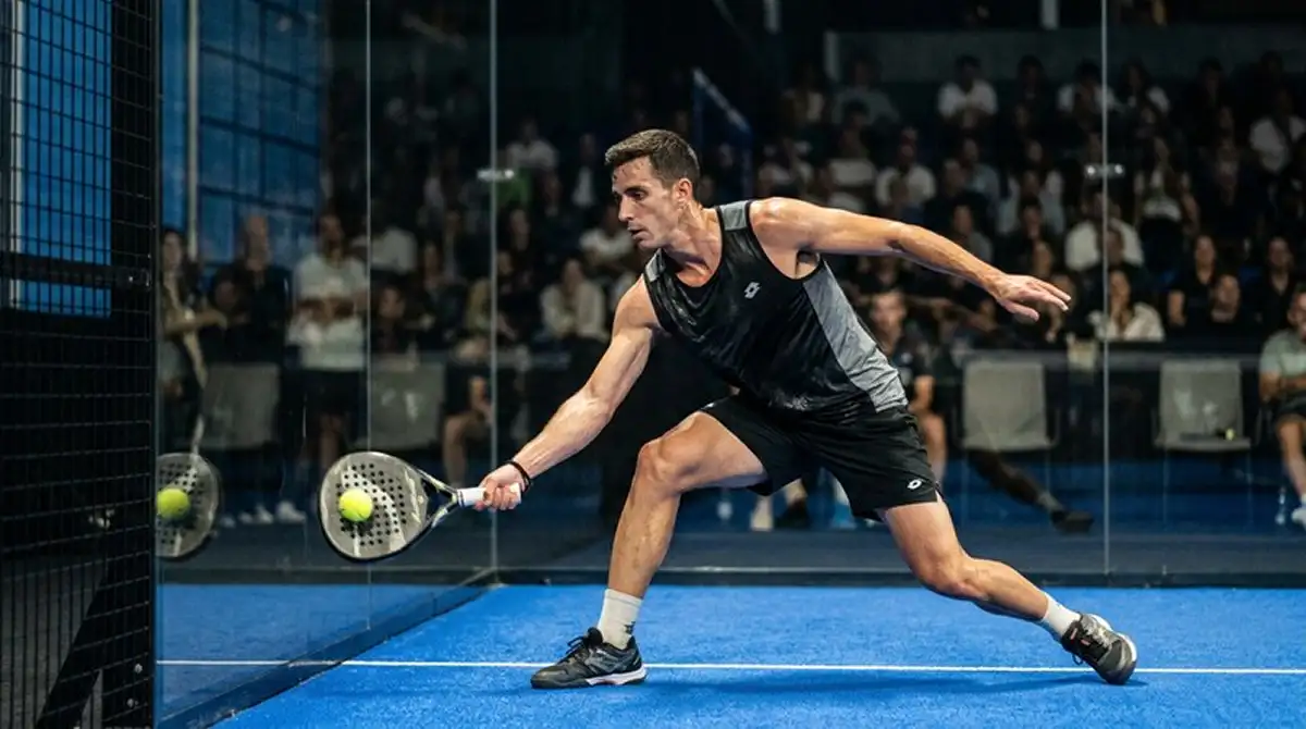 Giocatore di padel che colpisce di rovescio basso vicino alla parete in vetro durante un torneo indoor